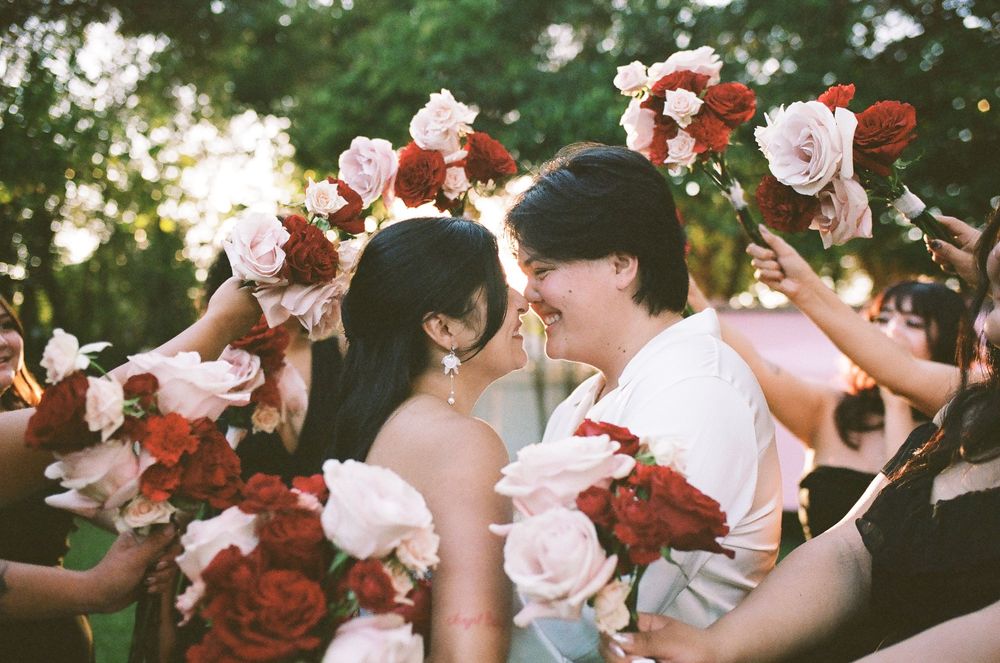 Two brides smile at each other while surrounded by guests holding red and white rose bouquets at an outdoor wedding ceremony.