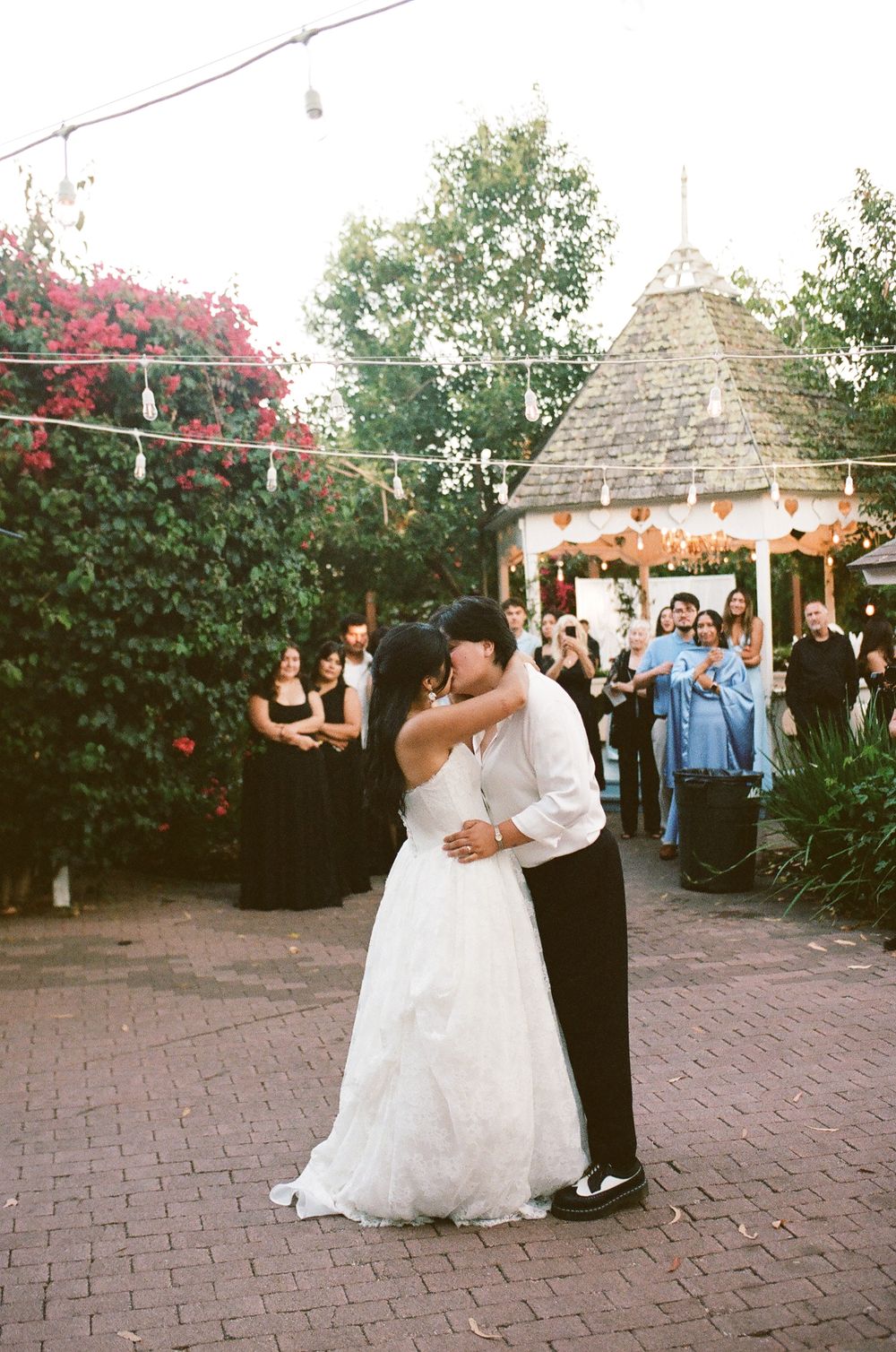 Bride in a white gown and groom in a white shirt share a first dance on a brick patio as wedding guests watch near a gazebo.
