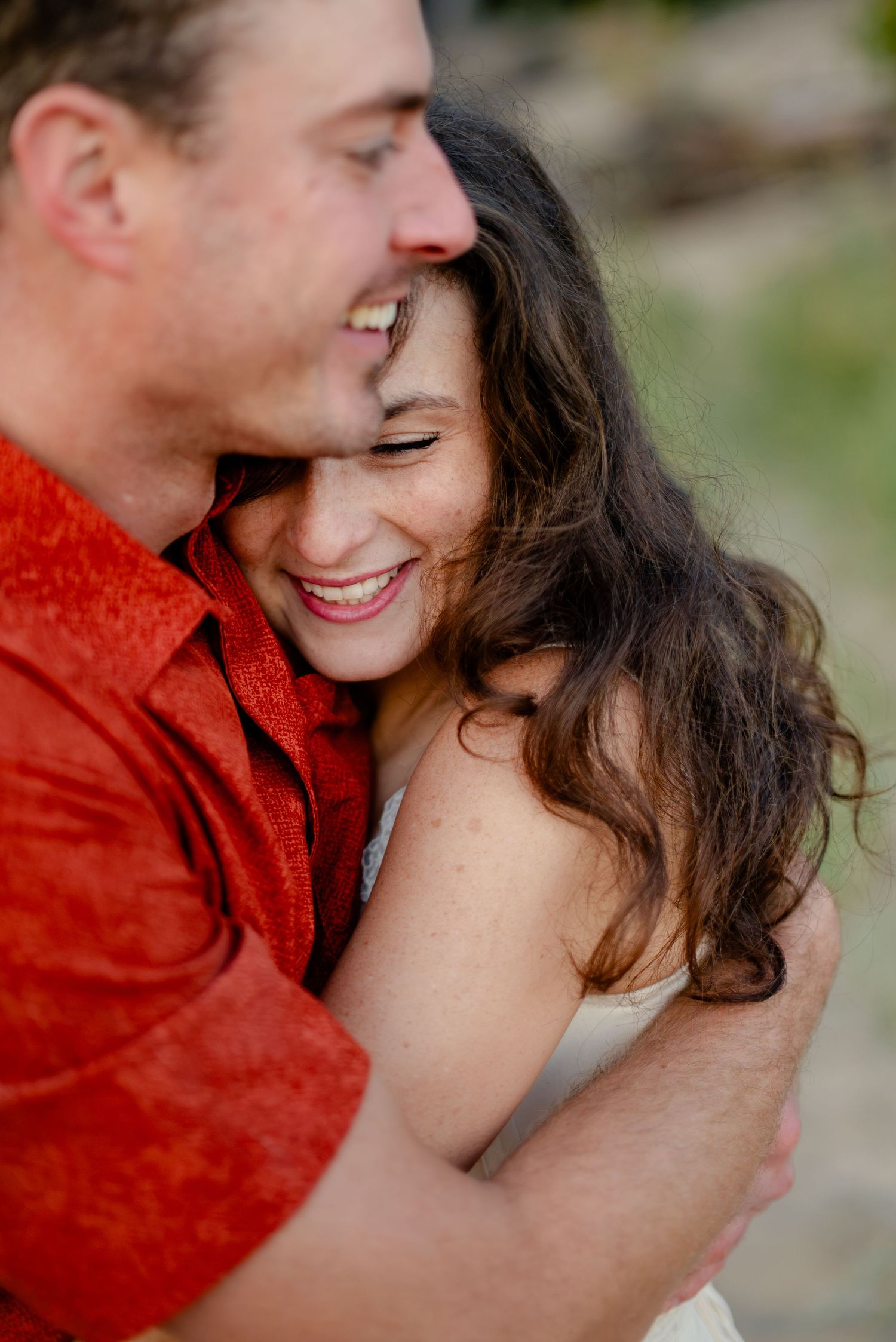 A joyful couple embraces warmly, the woman laughing as the man in a red shirt holds her close outdoors.