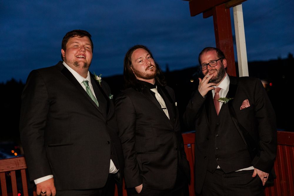 Three men in black suits smiling at an outdoor evening wedding reception.