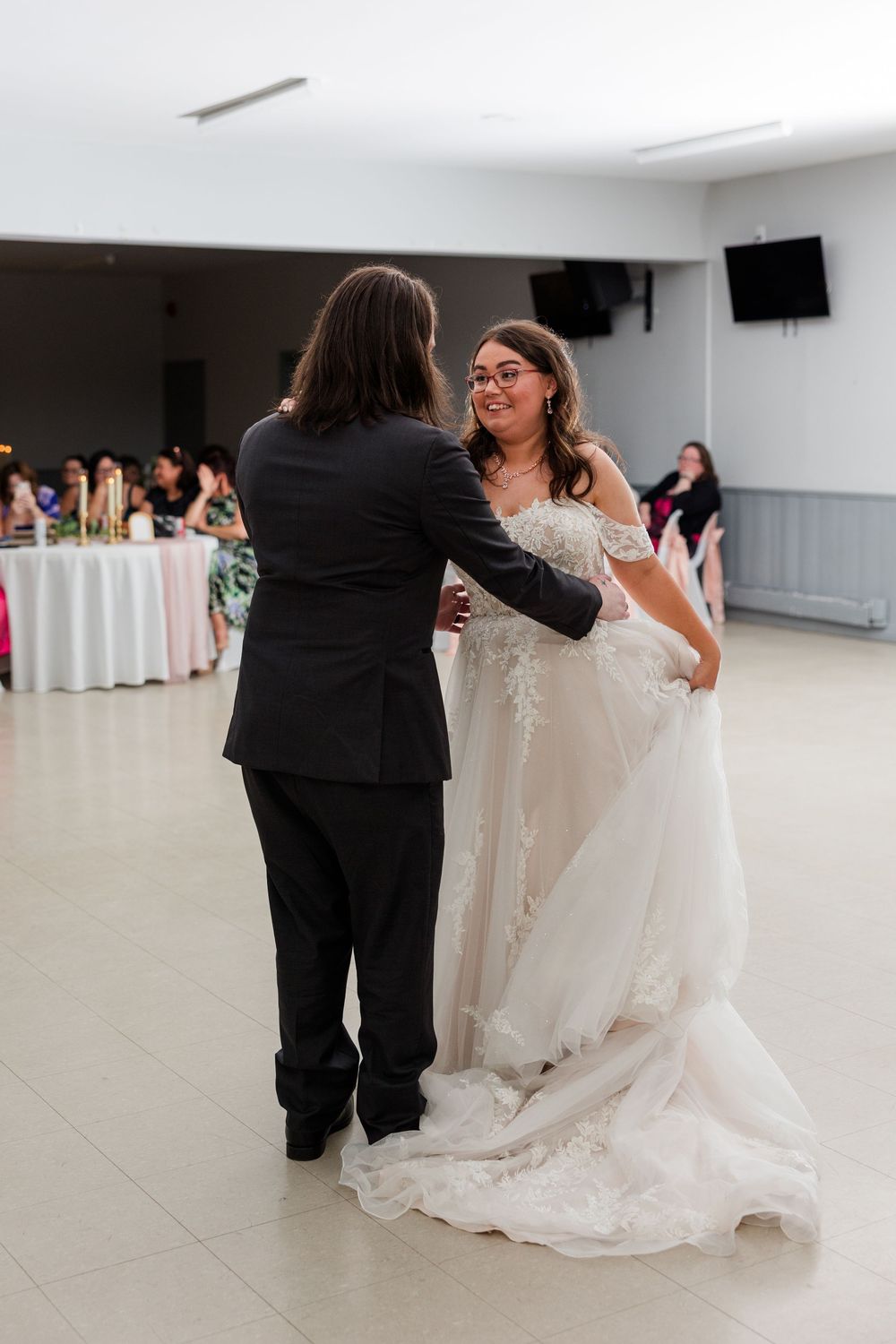Bride in lace wedding gown shares first dance with partner in black tuxedo at elegant wedding reception.