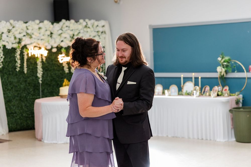 Groom in black suit shares mother-son dance with woman in purple dress at elegant wedding reception.