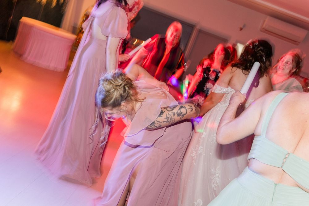 Bridesmaids in dusty rose gowns dancing energetically on a wedding reception dance floor with colorful lighting.