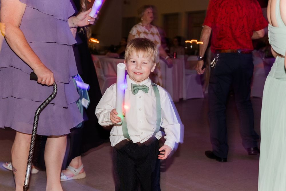 Young boy in suspenders and bow tie dancing with a glowing foam stick at a wedding reception.