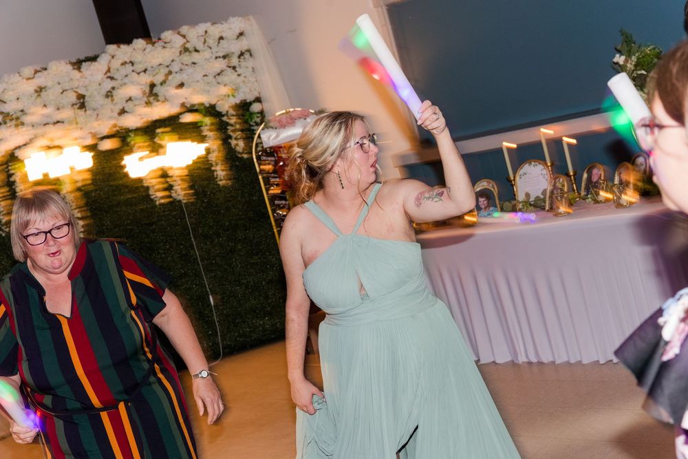 Women dancing at a wedding reception holding glowing light sticks in an elegant venue with floral decorations.