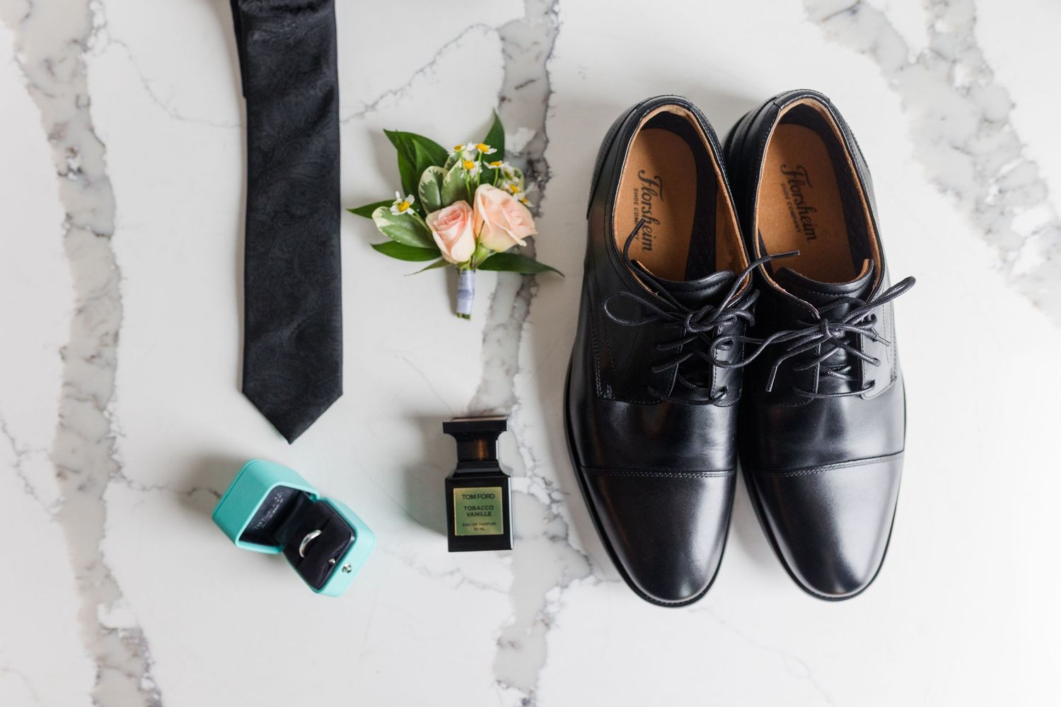 Black dress shoes, tie, boutonniere, cologne, and ring box arranged on marble surface for a groom's wedding day accessories.