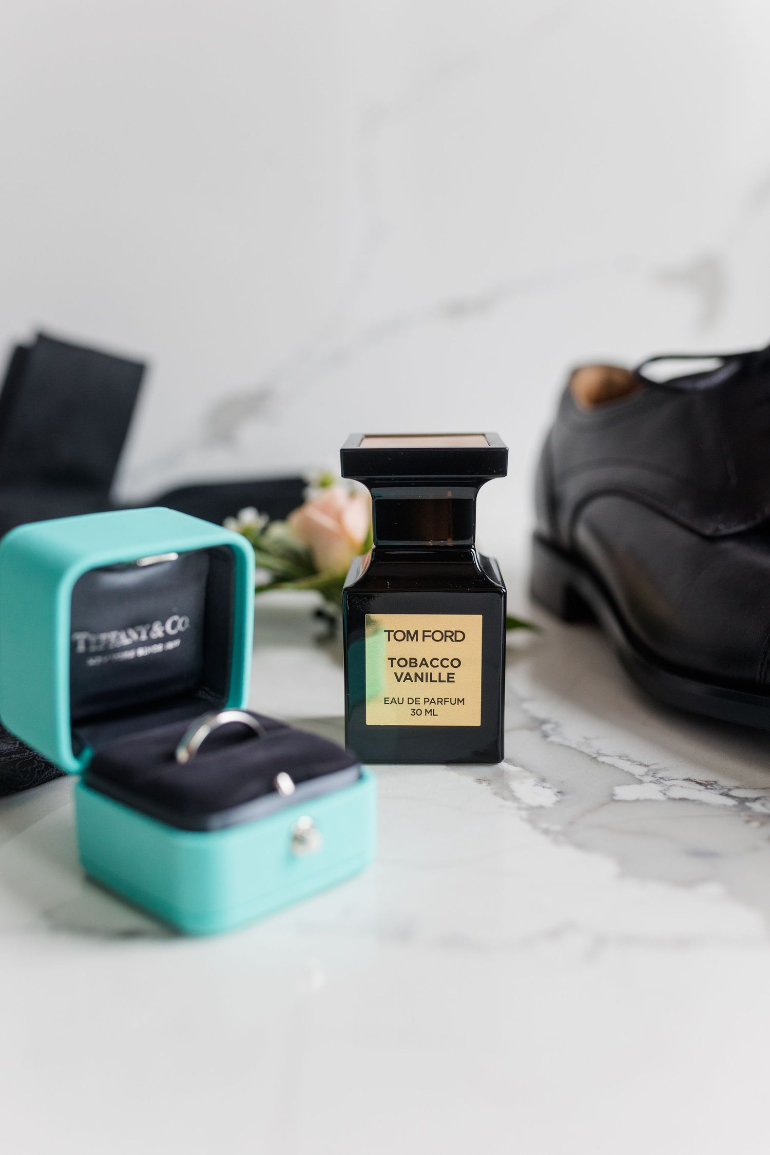 Tom Ford Tobacco Vanille perfume beside a Tiffany ring box, dress shoe, and rose on marble.