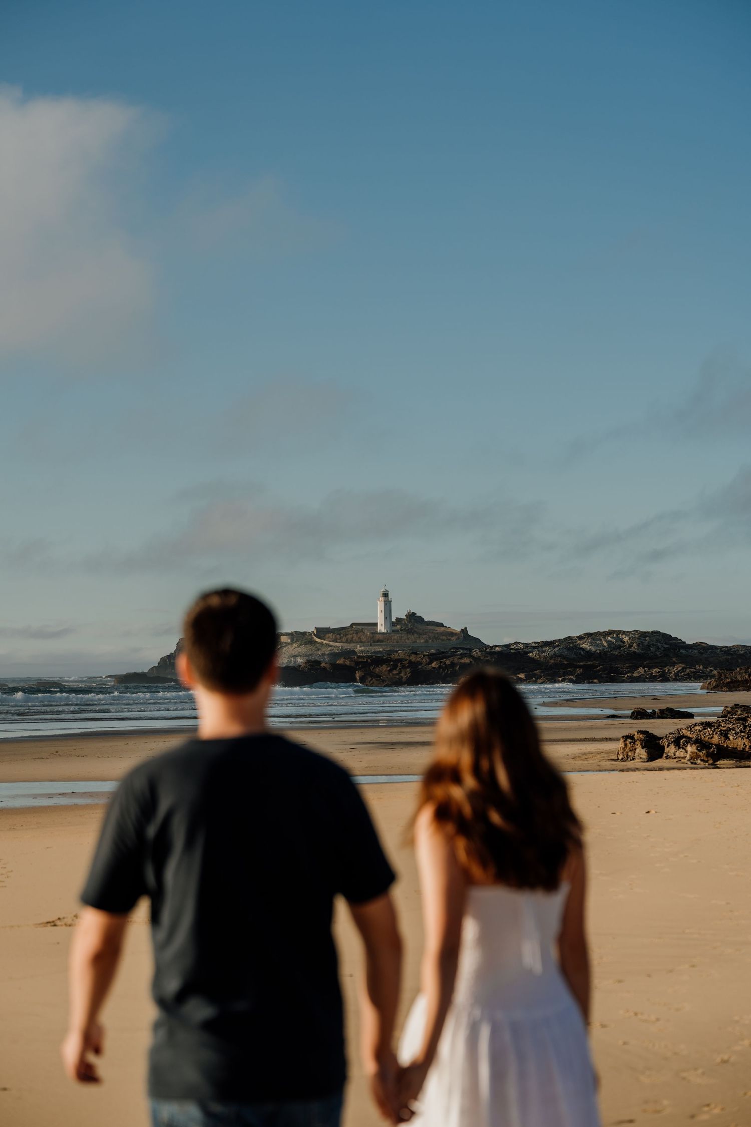 Couple holding hands on sandy beach facing Godrevy lighthouse in St Ives bay.