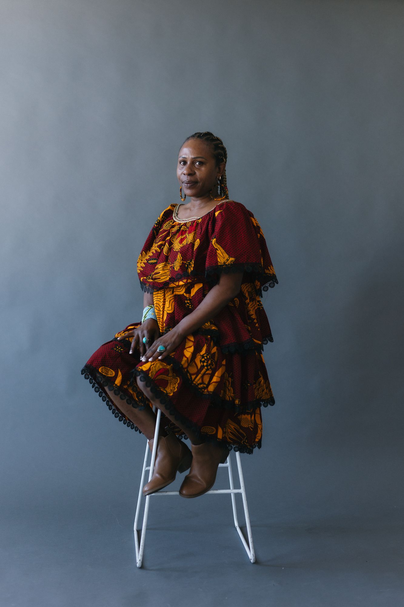 Woman in colorful African print dress seated on white stool against gray studio background.