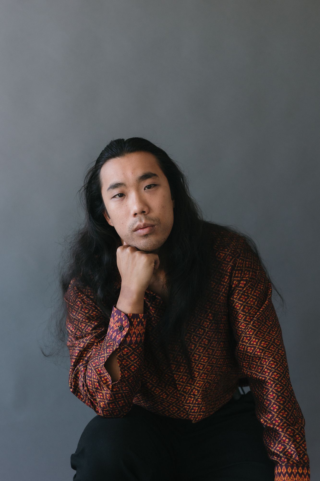 Asian man with long black hair in patterned red shirt poses thoughtfully against gray background.