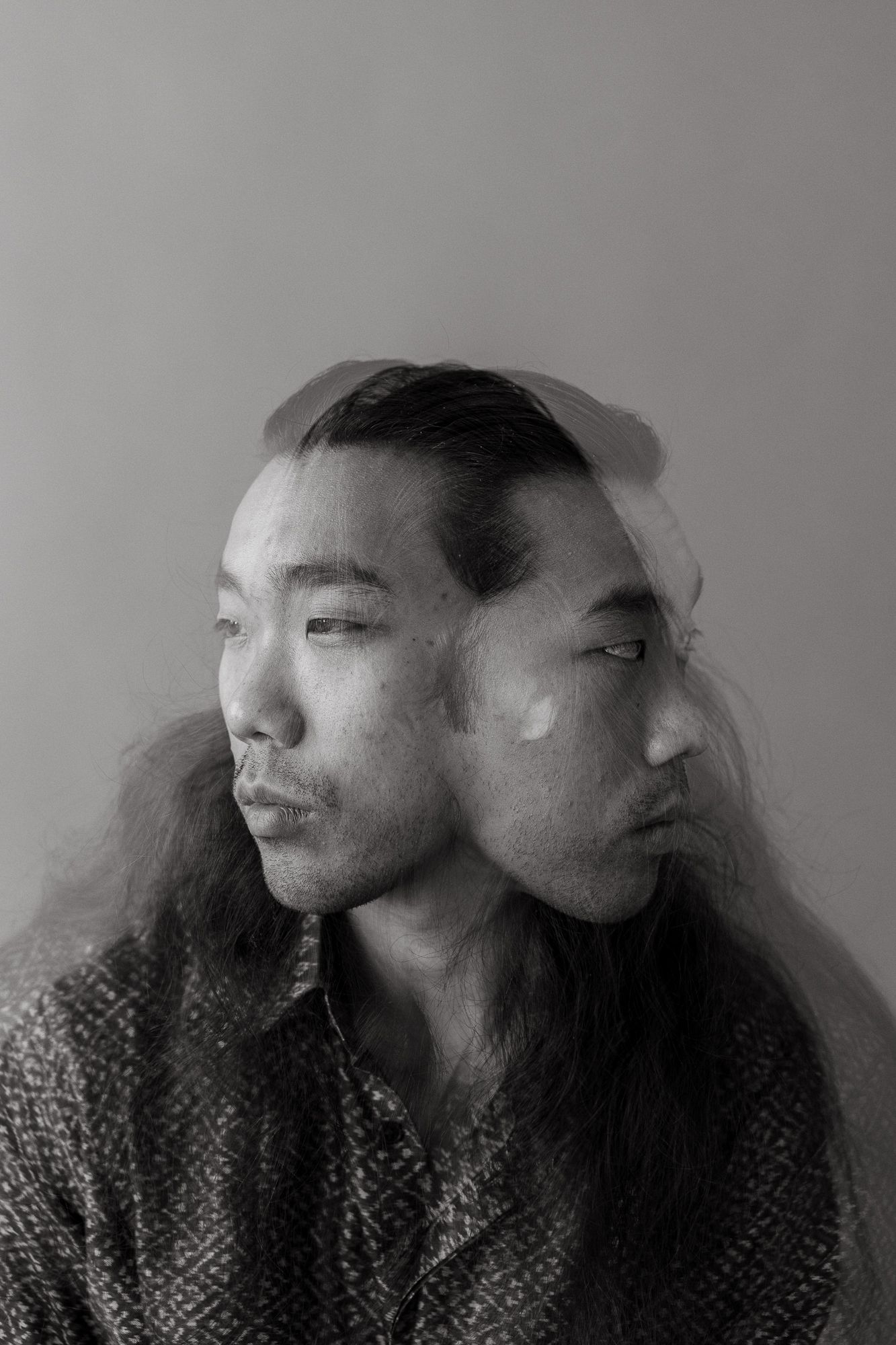 Black and white double exposure portrait of an Asian man with long hair, creating a surreal multi-face effect.