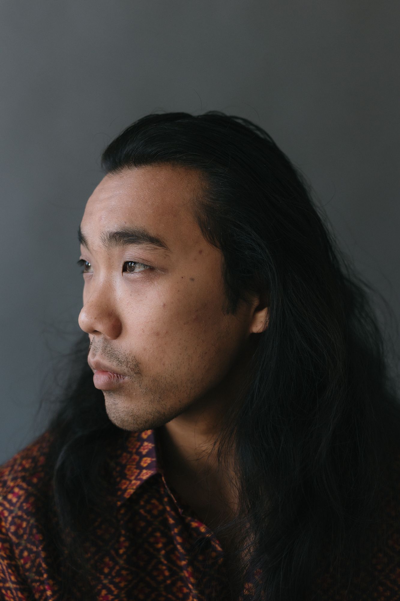 Profile portrait of a young Asian man with long black hair wearing a patterned red shirt against a gray background.