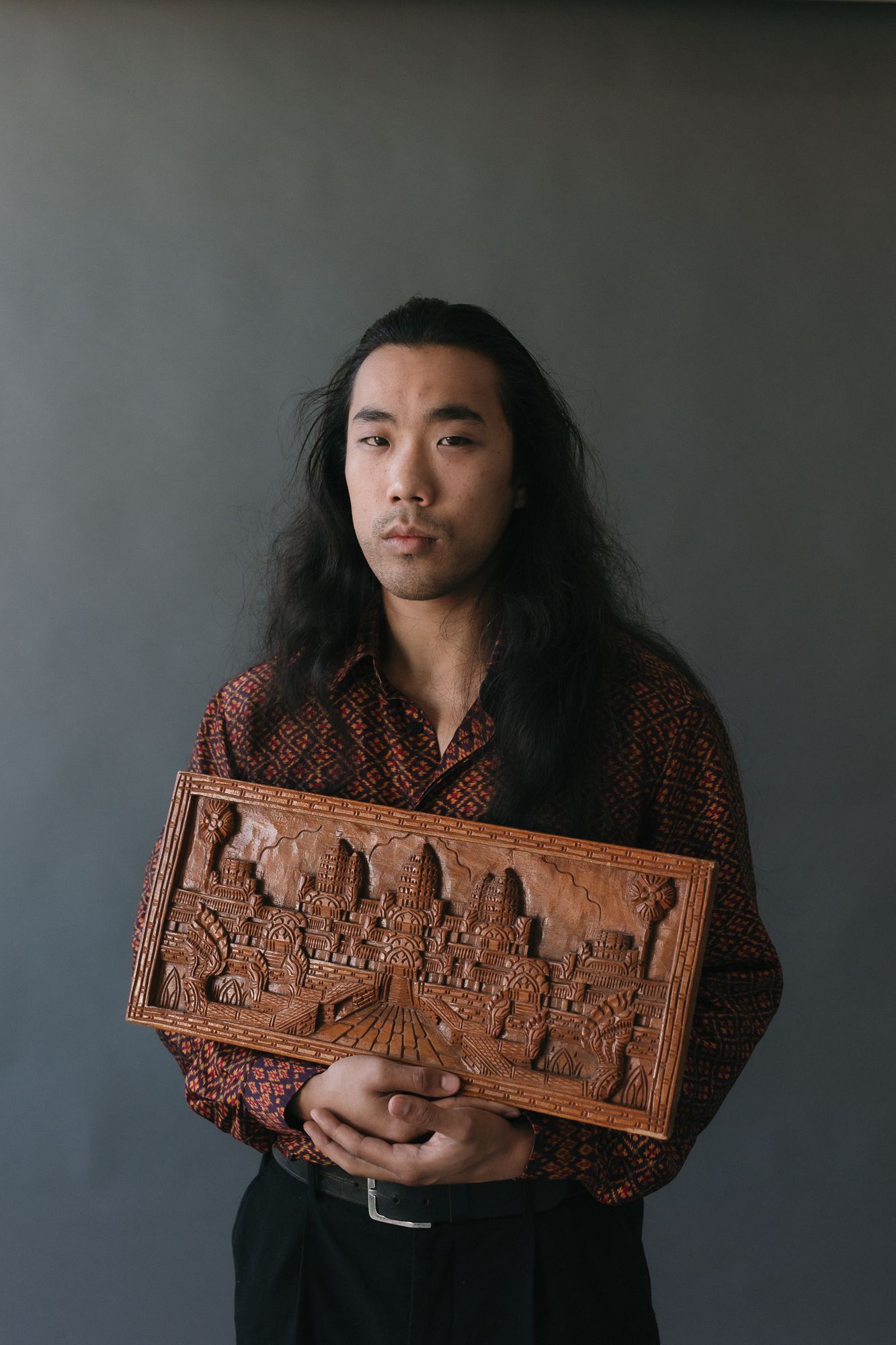 Young man with long black hair holding an ornate wooden carved panel, wearing a batik shirt against gray background.