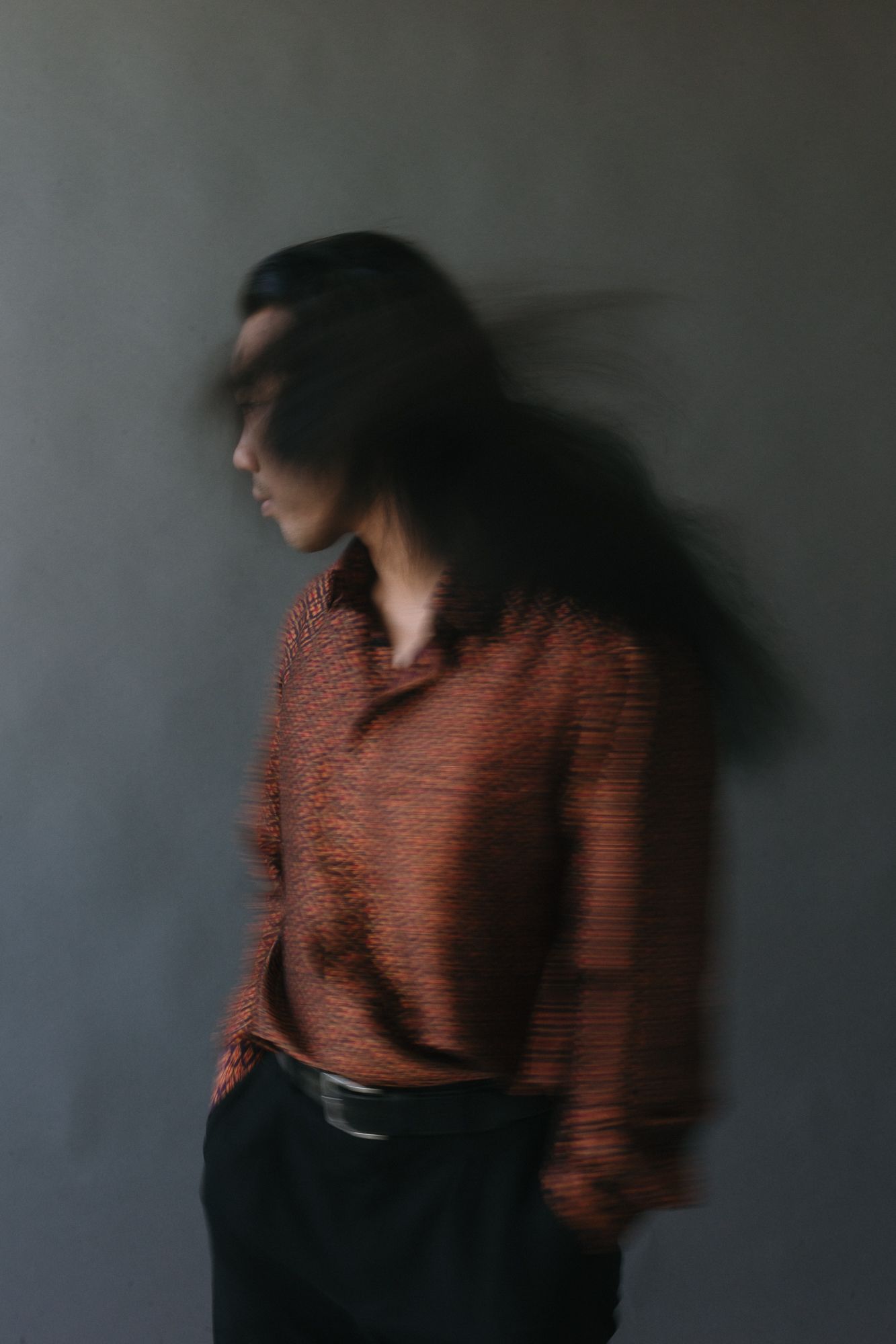 Blurred motion portrait of a person with long dark hair wearing a red textured shirt against a gray wall.