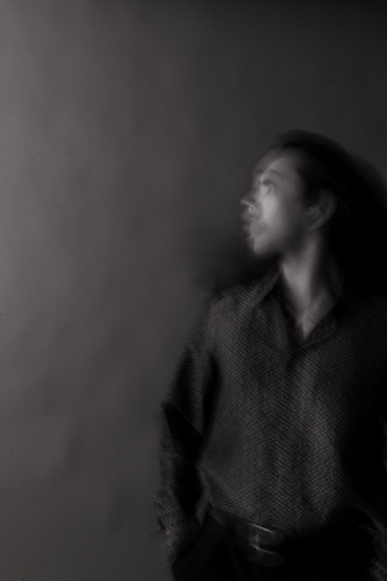 Black and white blurred portrait of a person with long hair in a textured shirt gazing upward dramatically.