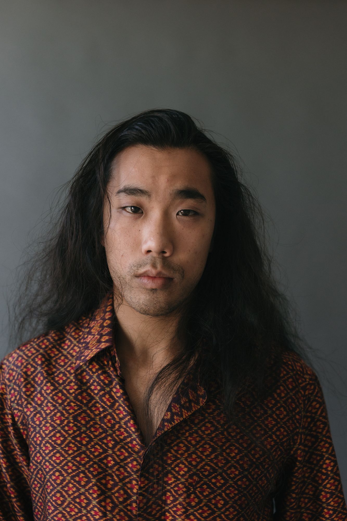 Young Asian man with long black hair wearing a patterned red shirt, posing against a gray background.