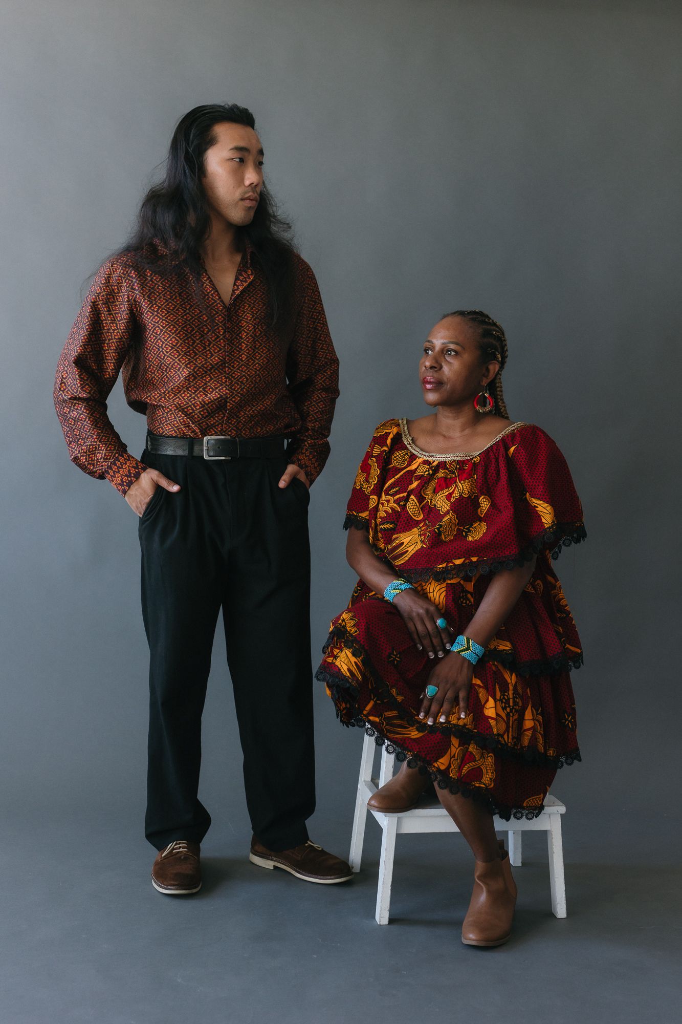 A young man with long hair and a woman in colorful African print dress pose together in a studio portrait.