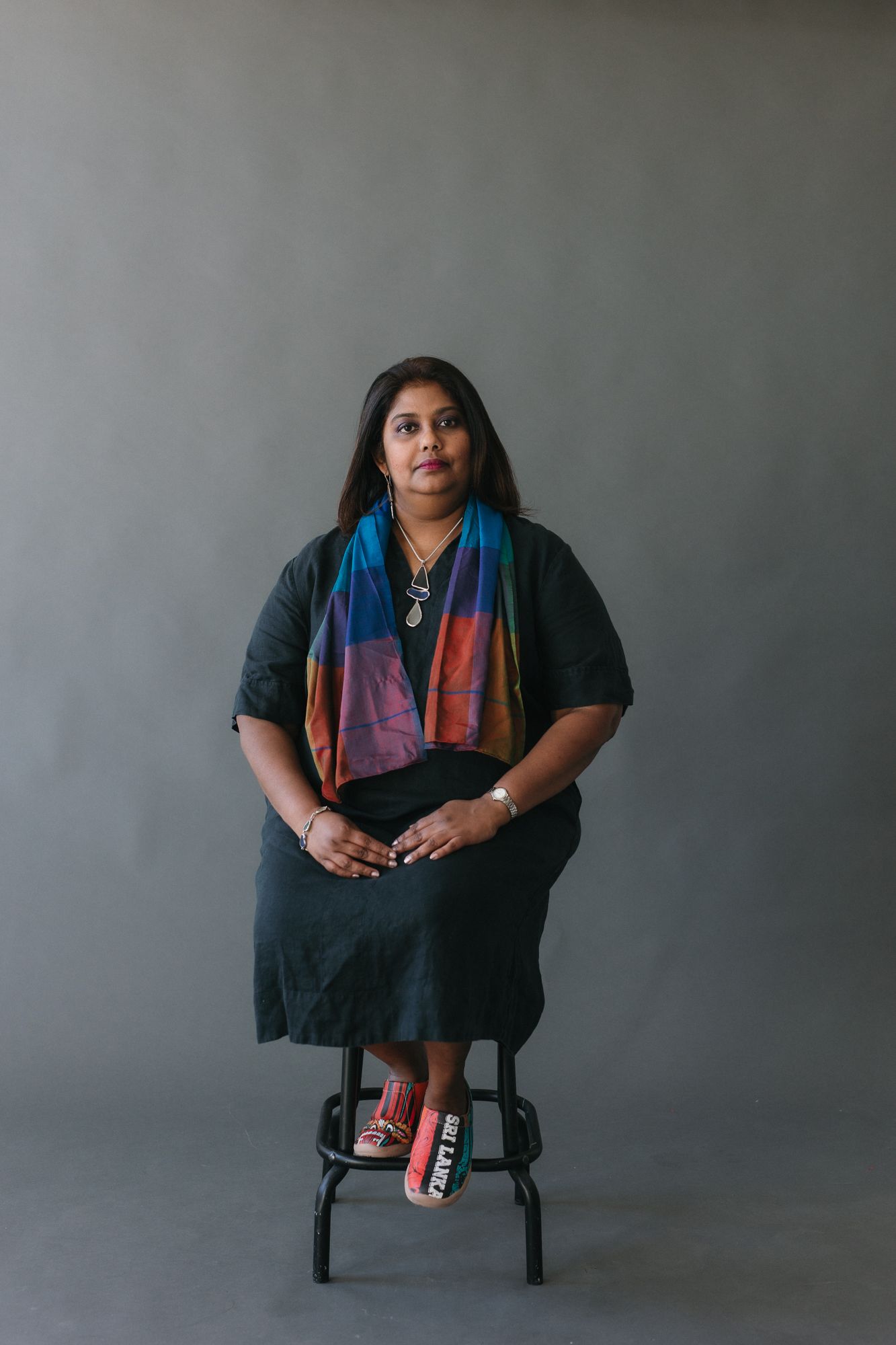 Woman in black dress with colorful scarf sits on stool against gray background in studio portrait.