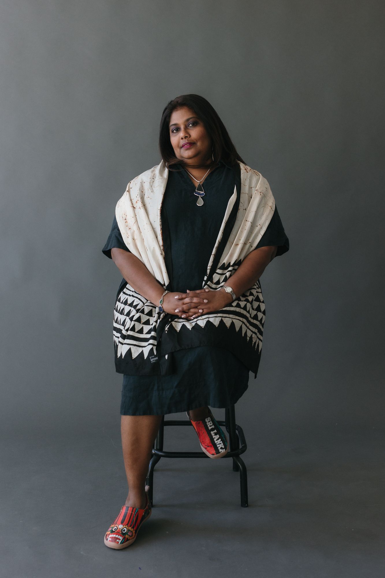 A woman in a black dress and patterned shawl sits on a stool against a gray background.