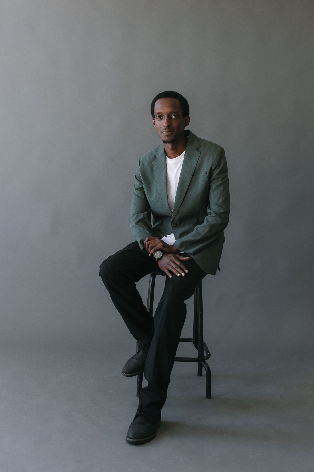 A man in a green blazer and black pants sits on a stool against a gray background.