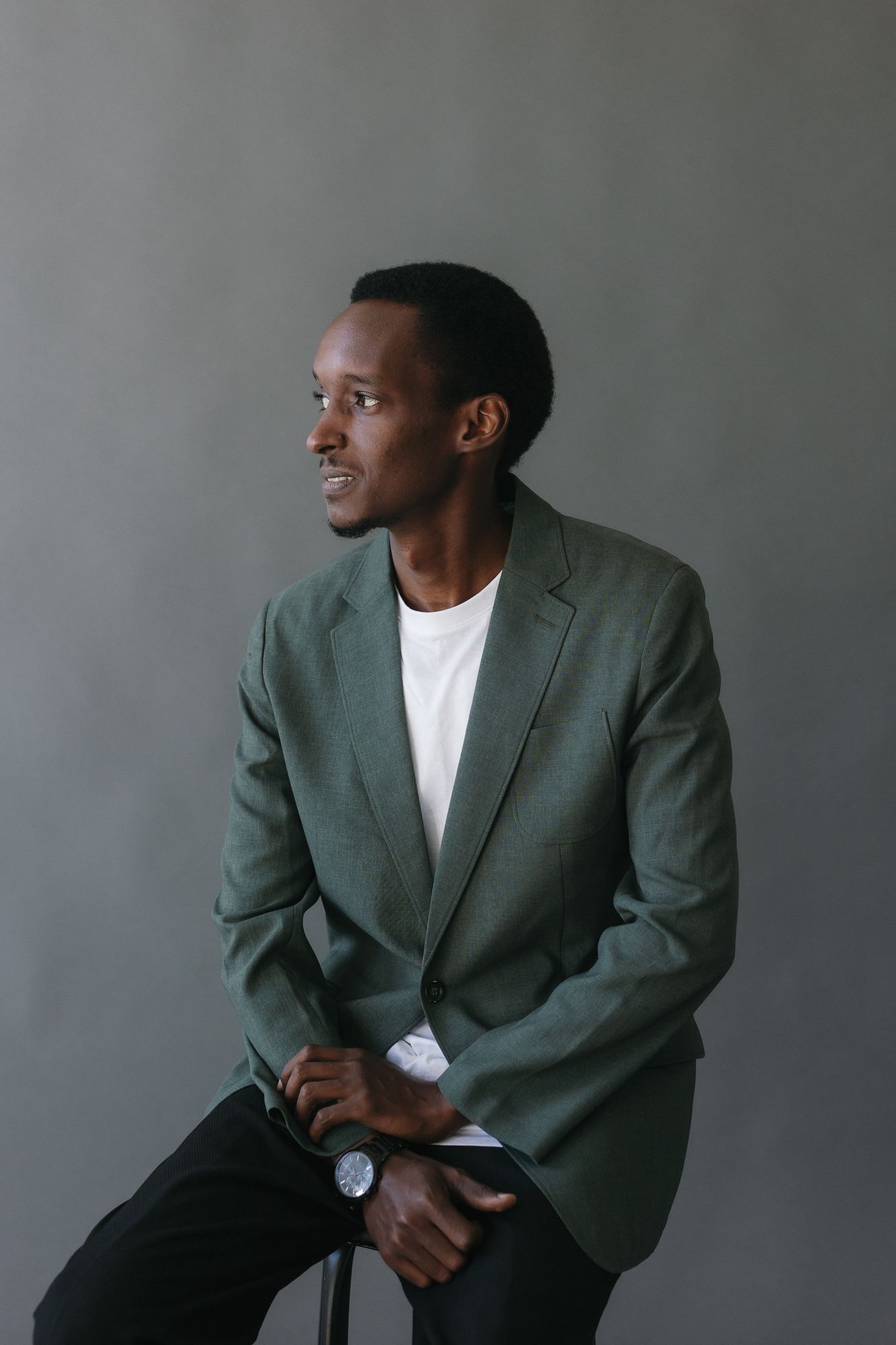 A young Black man in a green blazer and white shirt sits confidently, wearing a watch, against a gray background.