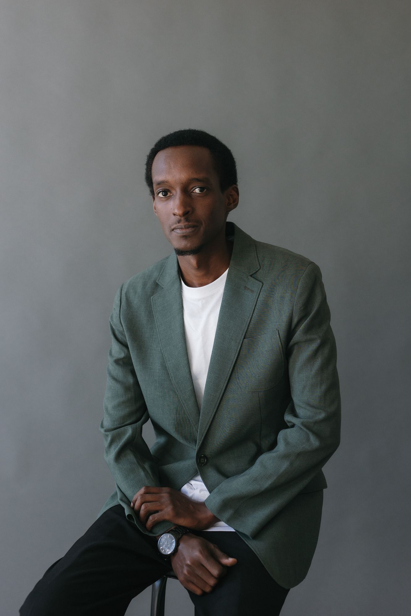 Man in green blazer and white shirt sits against gray background, looking at camera with a watch on his wrist.