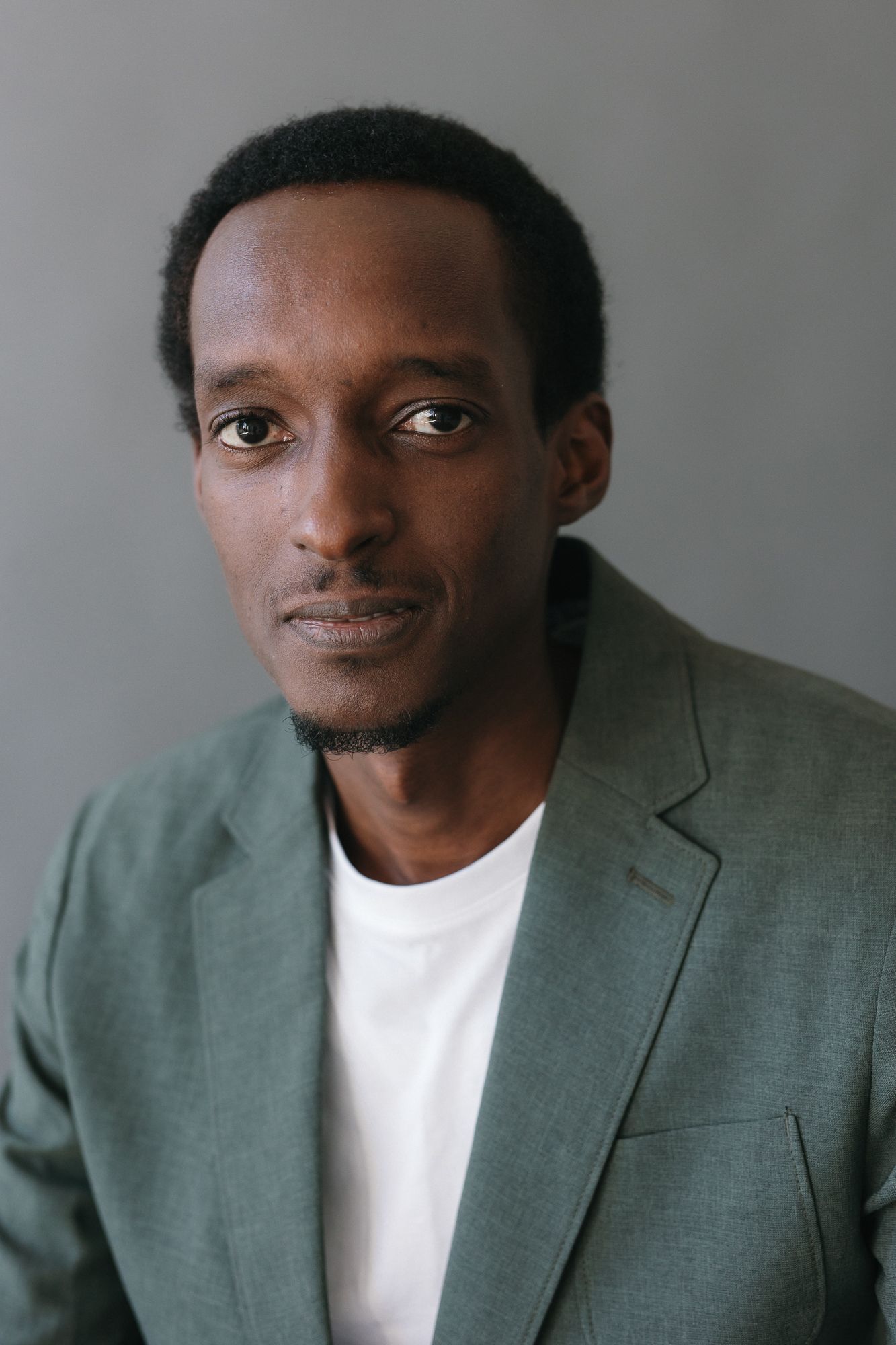 Young Black man in green blazer and white t-shirt poses against gray background in professional headshot.
