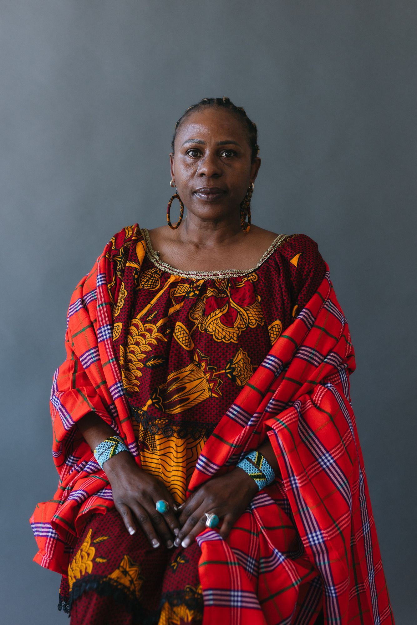 A dignified woman in vibrant red plaid and African print dress with turquoise rings poses against gray background.