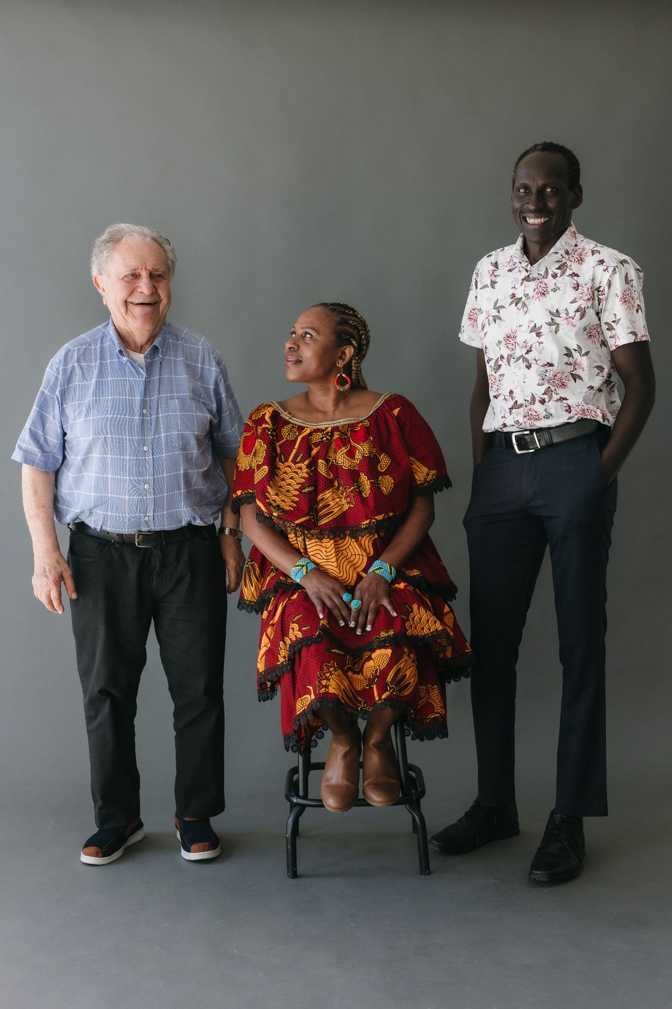 Three people posing together in a studio, woman in colorful African dress seated between two standing men smiling.
