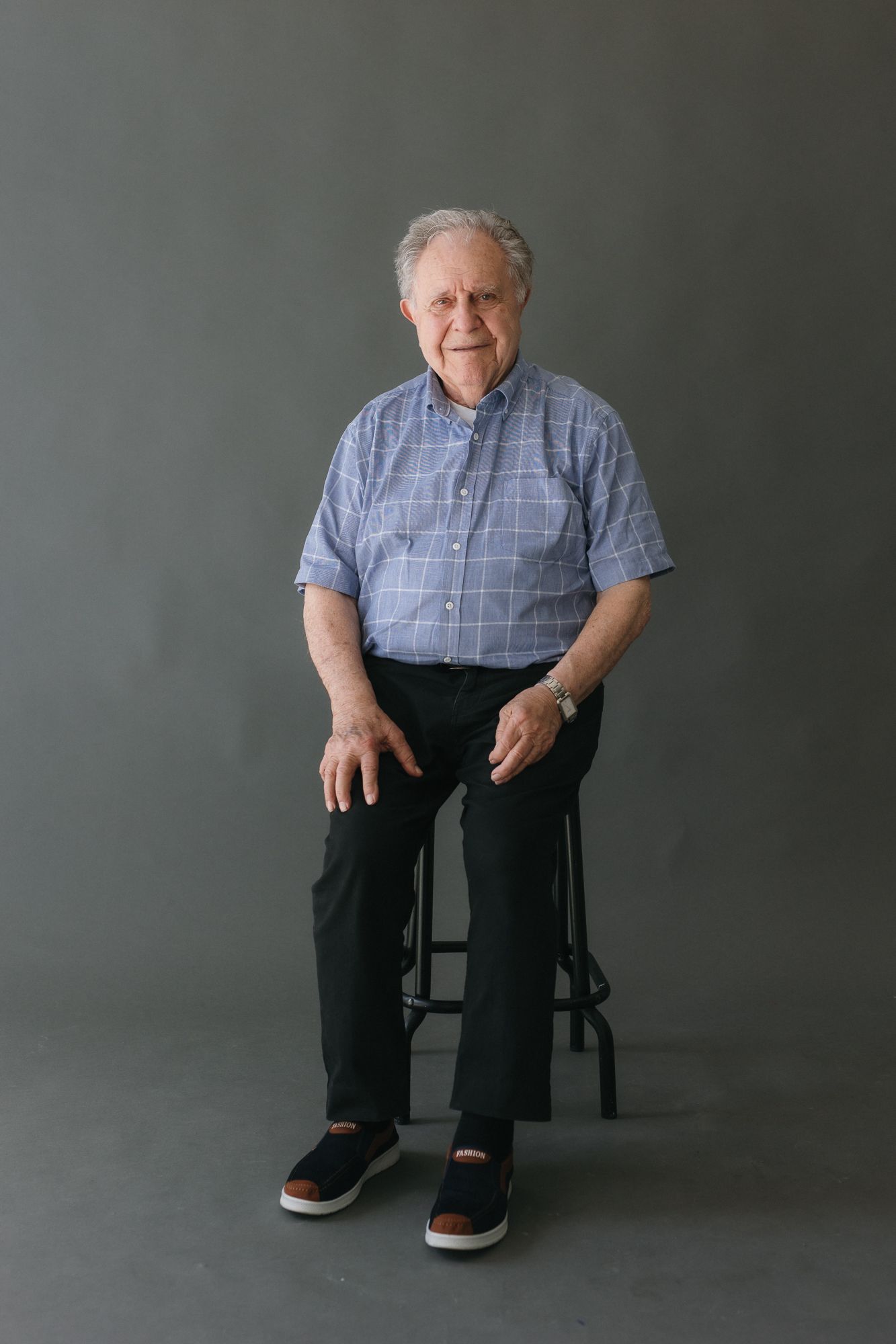 Elderly man in plaid shirt and black pants sitting on a stool against a gray studio background.