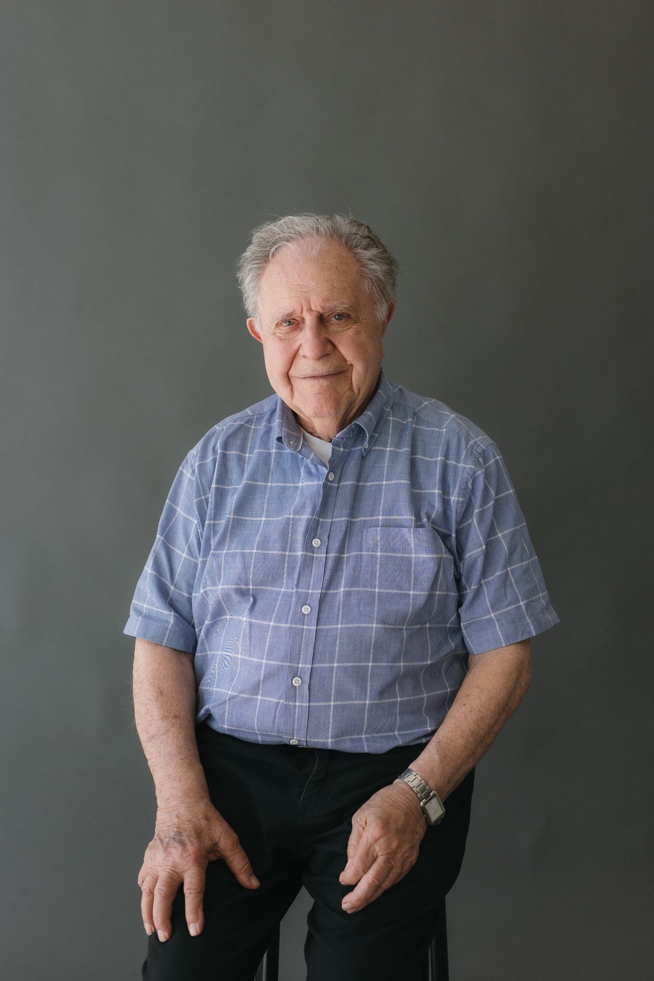 Elderly man in blue plaid short-sleeve shirt sitting against gray background, wearing a watch, looking at camera.