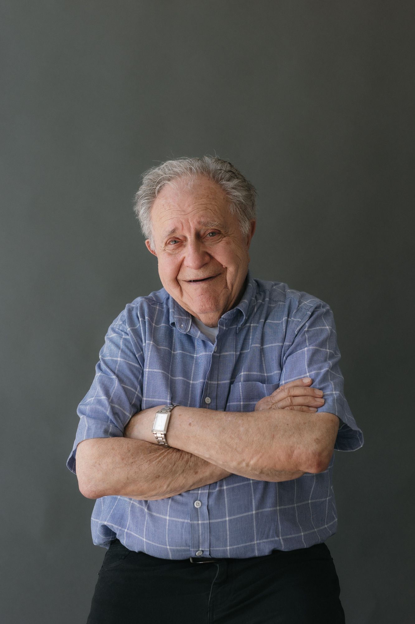 Elderly man with gray hair smiling, arms crossed, wearing a blue plaid shirt and watch against a gray background.