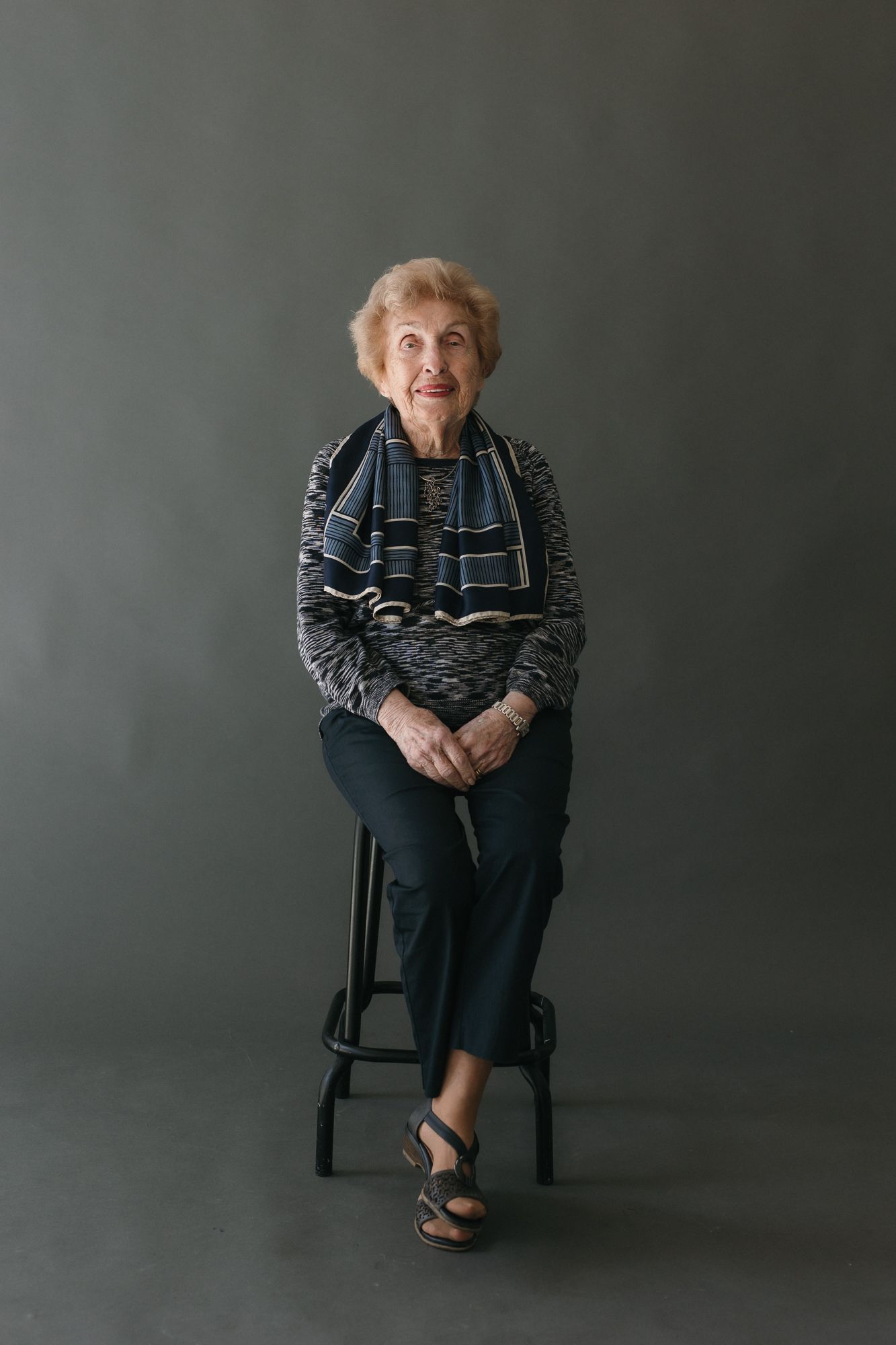 Elderly woman sitting on a stool wearing a striped scarf and dark pants, posing for a portrait against a gray background.