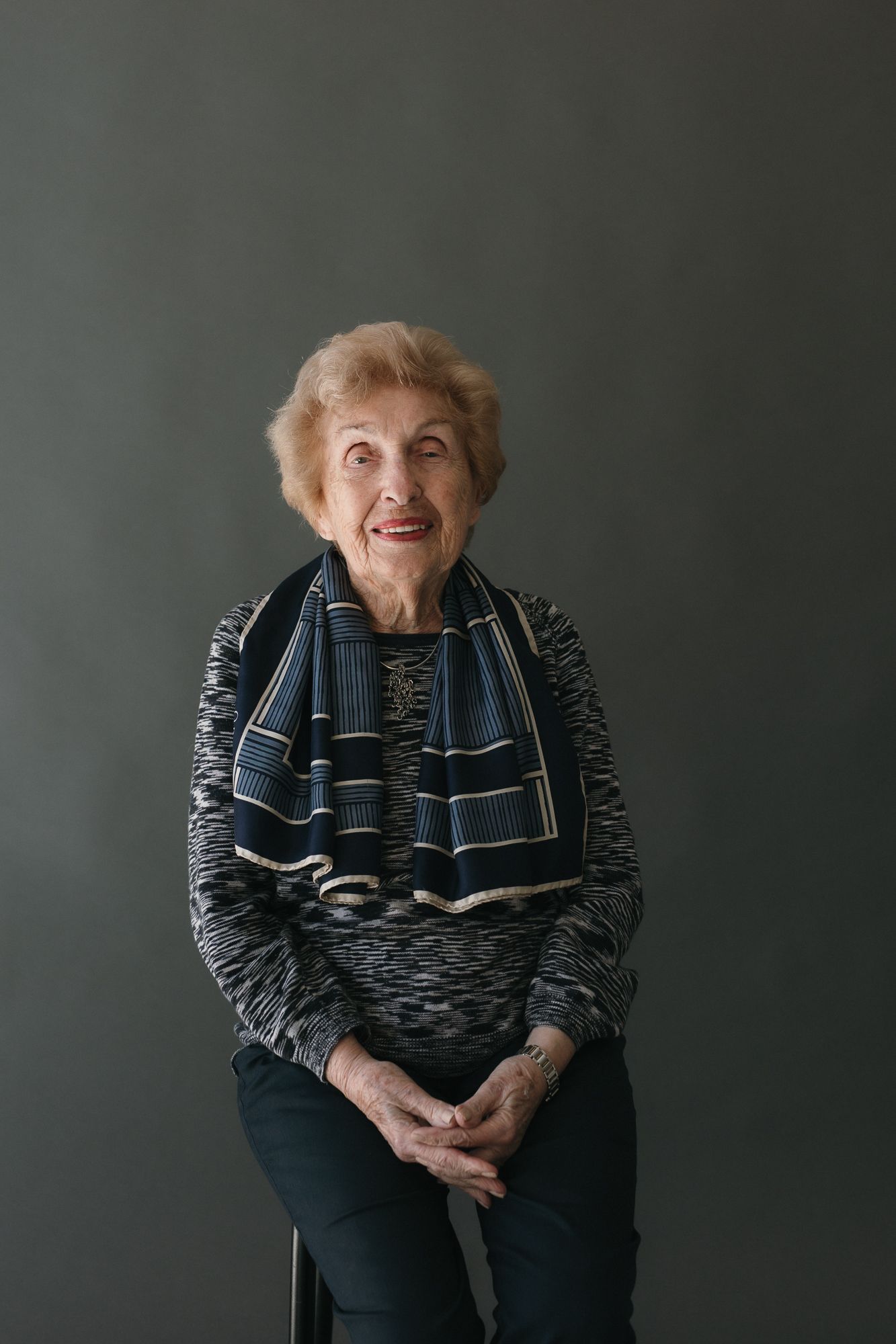 Elderly woman with short blonde hair wearing a striped scarf and patterned sweater, seated and smiling against a gray background.