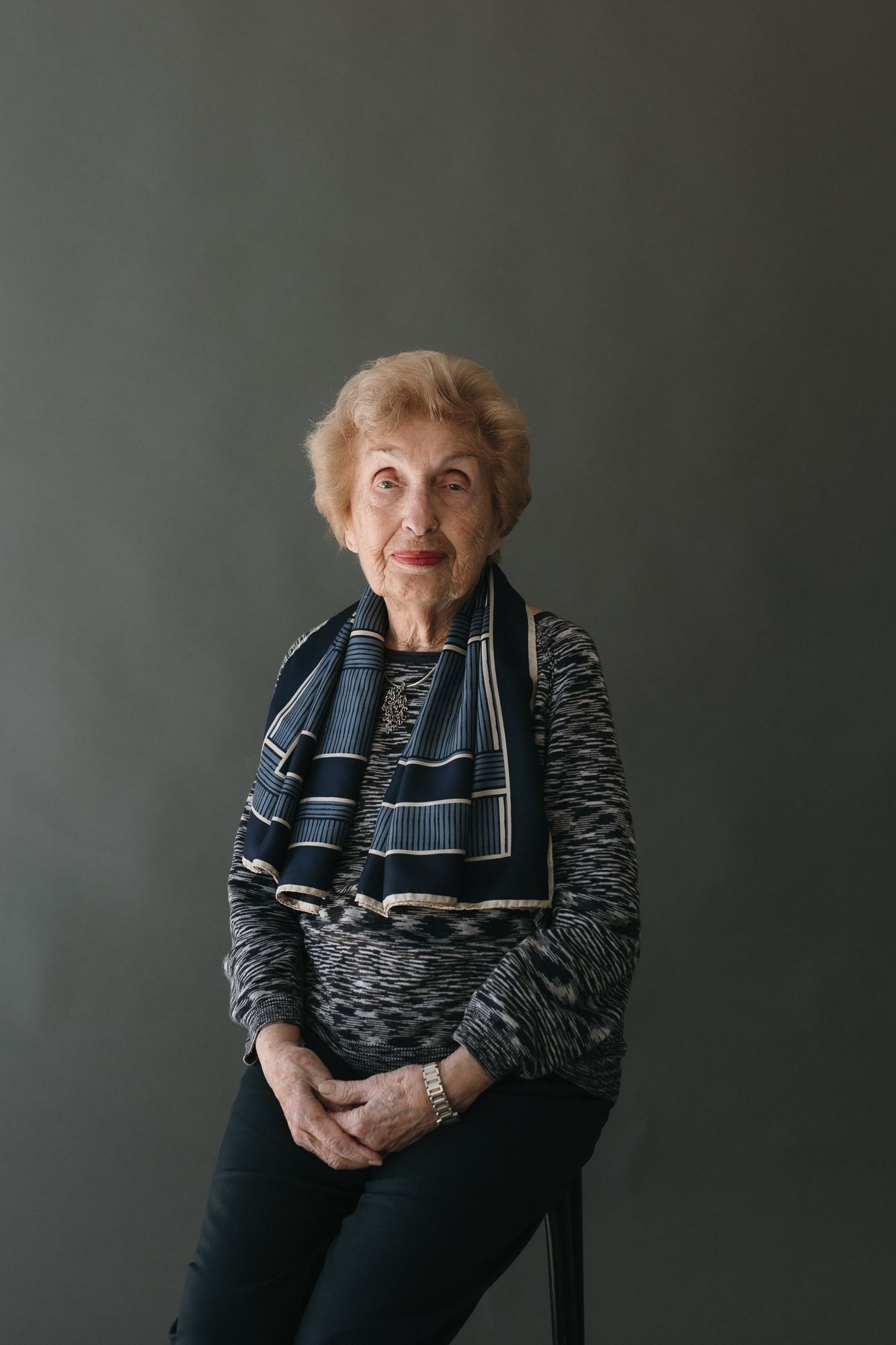 Elderly woman with short blonde hair wearing a striped scarf and patterned top, seated against a gray background.