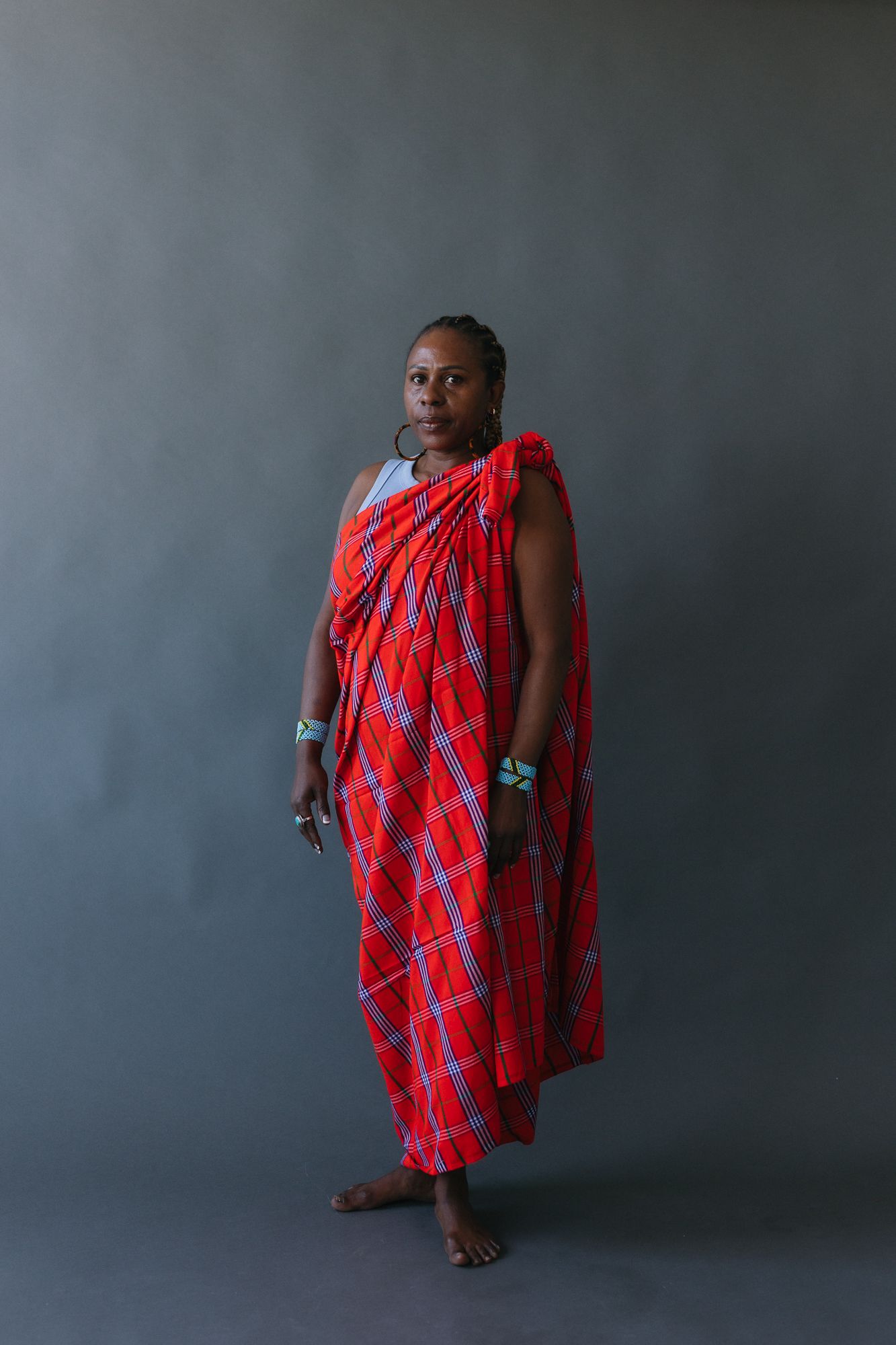 Woman in traditional red plaid Maasai shuka dress with turquoise beaded jewelry poses against gray background.