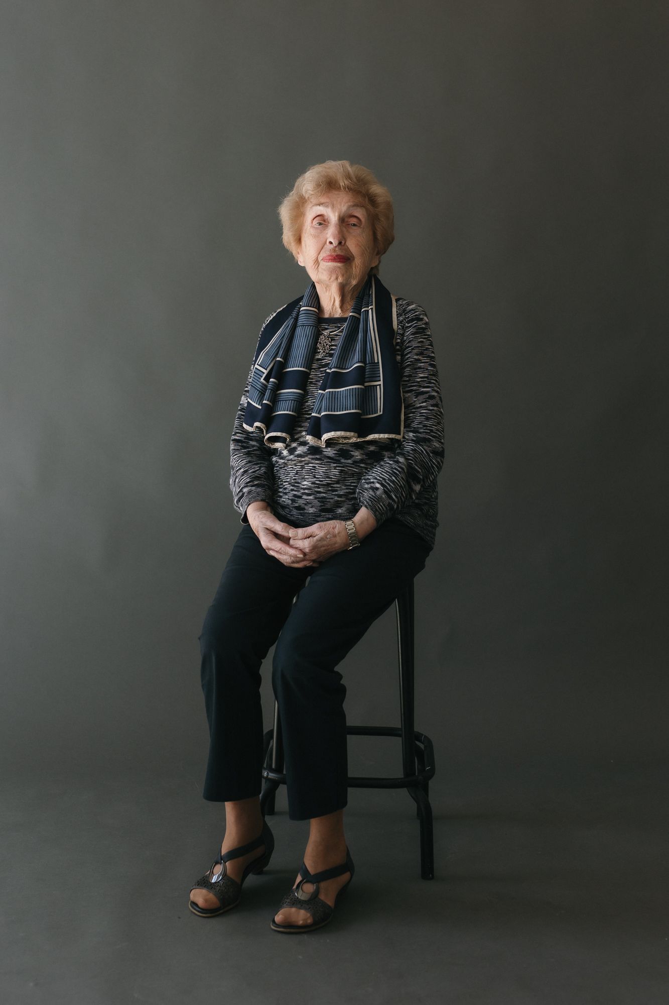 Elderly woman sitting on a stool wearing black pants and a striped scarf against a gray background.