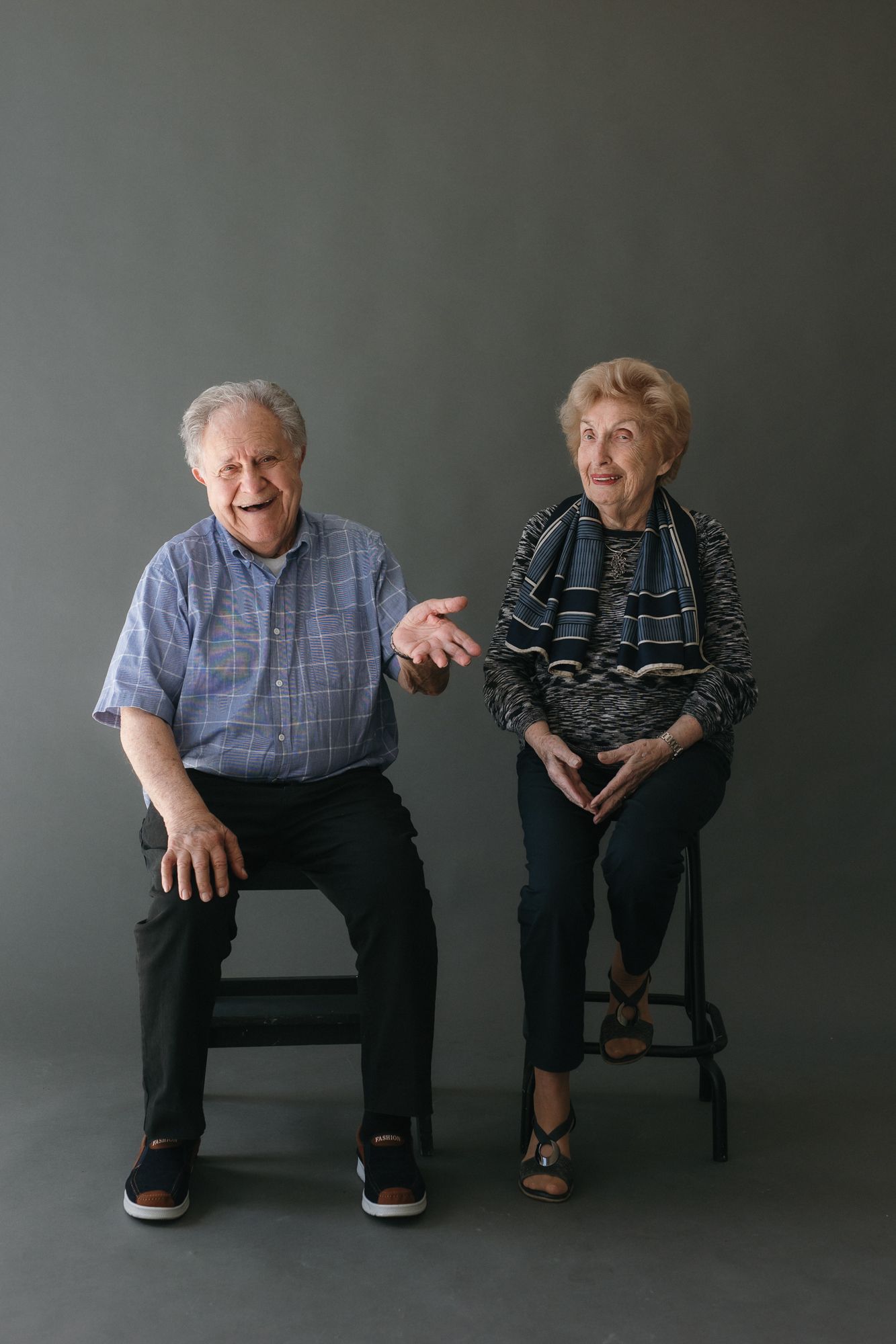 Two elderly people sitting on stools smiling in a studio portrait against a gray background.