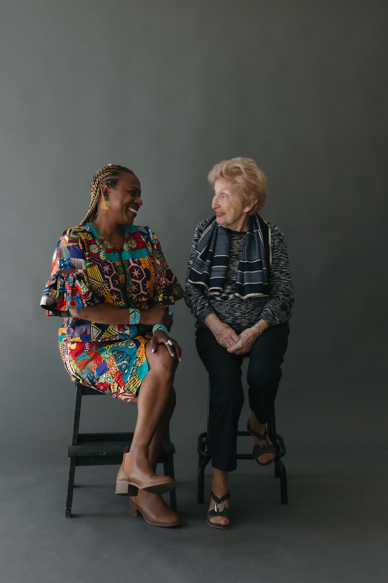 Two women of different ages sit on stools laughing together, one in colorful dress, one in striped top.