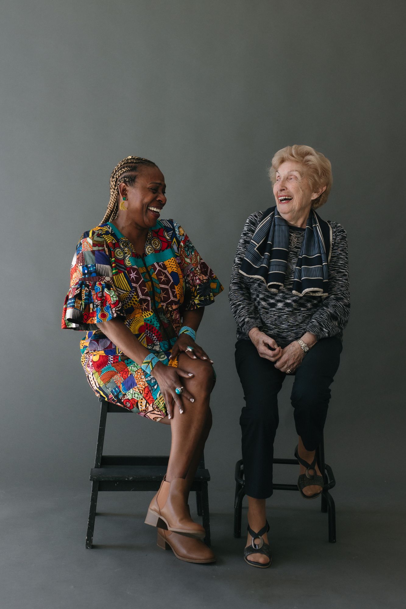 Two women sitting on stools laughing together, one in colorful African print dress, one in striped top, against gray background.