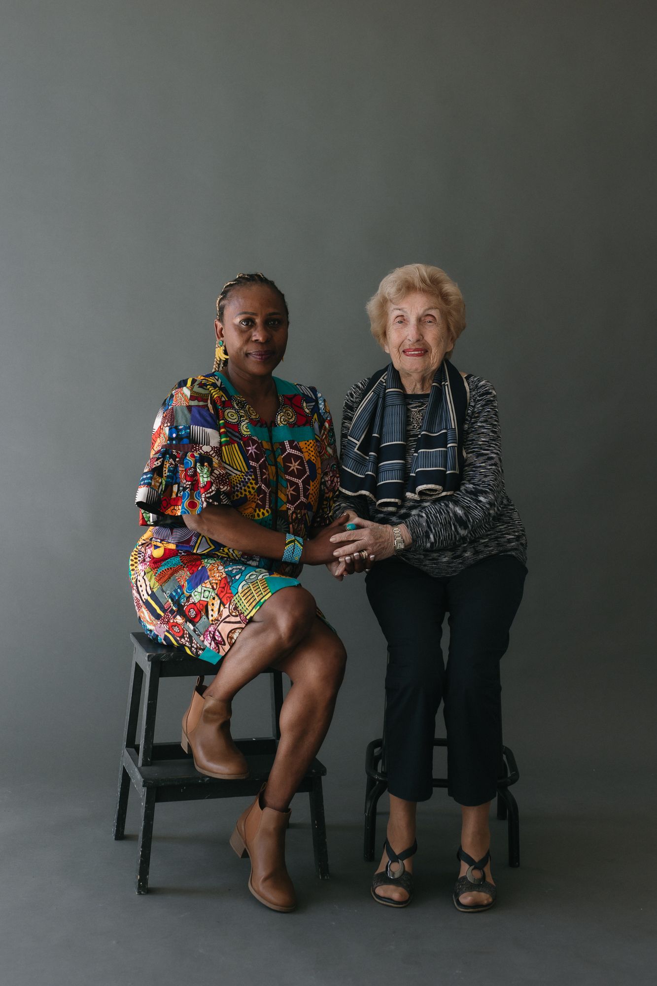 Two women of different generations sit together on stools, smiling, against a gray studio background.