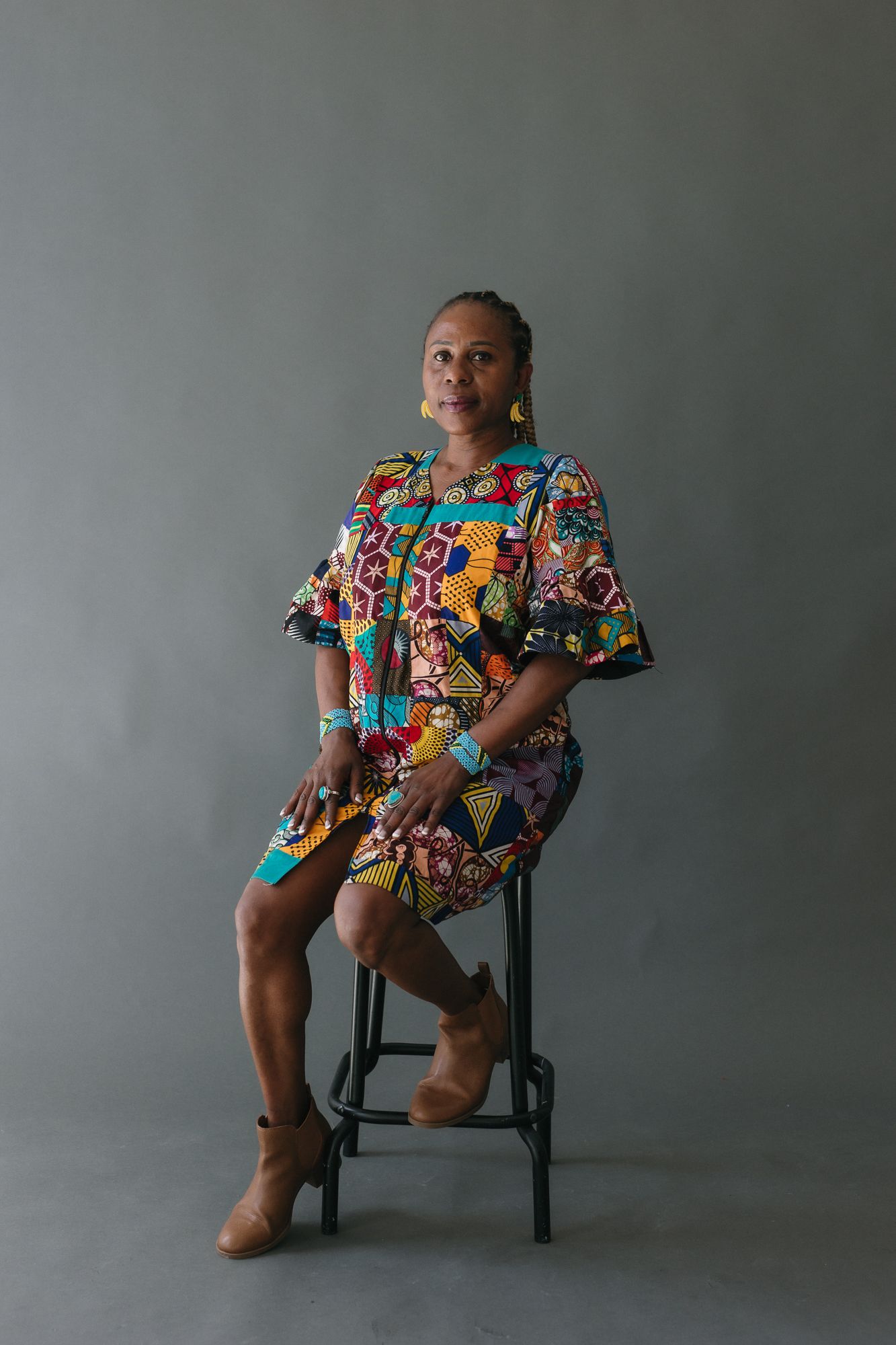 Woman in colorful African print dress seated on stool against gray background.