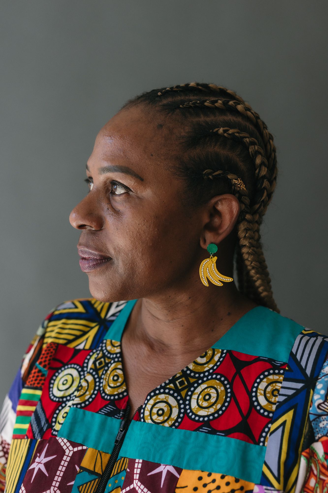 African woman in colorful traditional print top with braided hair and banana earrings, looking sideways against gray background.