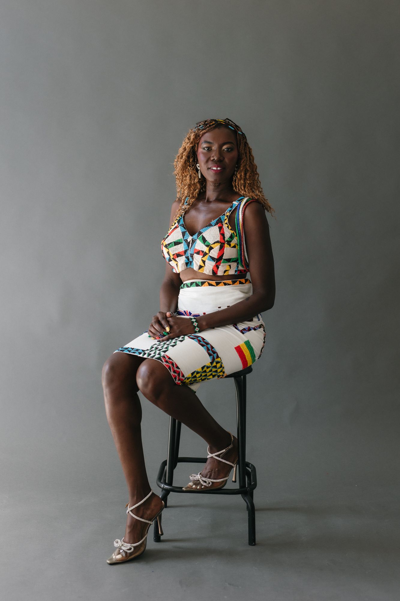 Young woman in white African patterned two-piece outfit seated on stool.