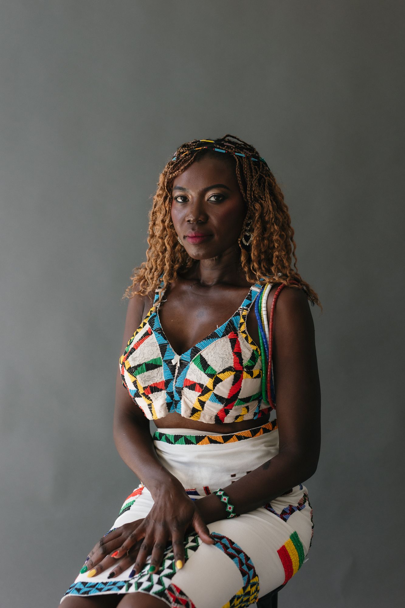 Woman in colorful African print crop top and skirt with braided hair sits confidently against gray background.