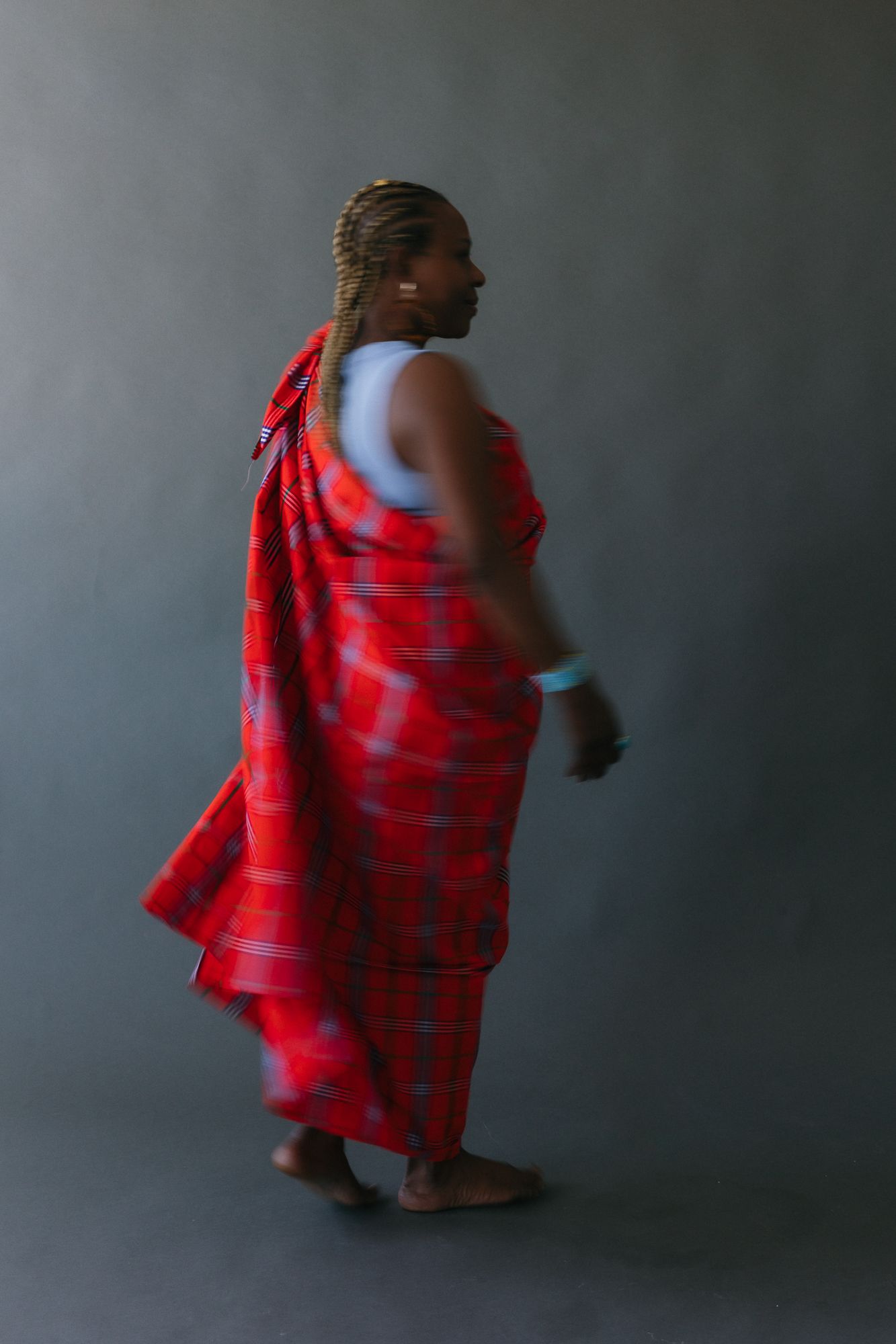 A Maasai man wearing traditional red plaid shuka drape stands barefoot against a gray background.