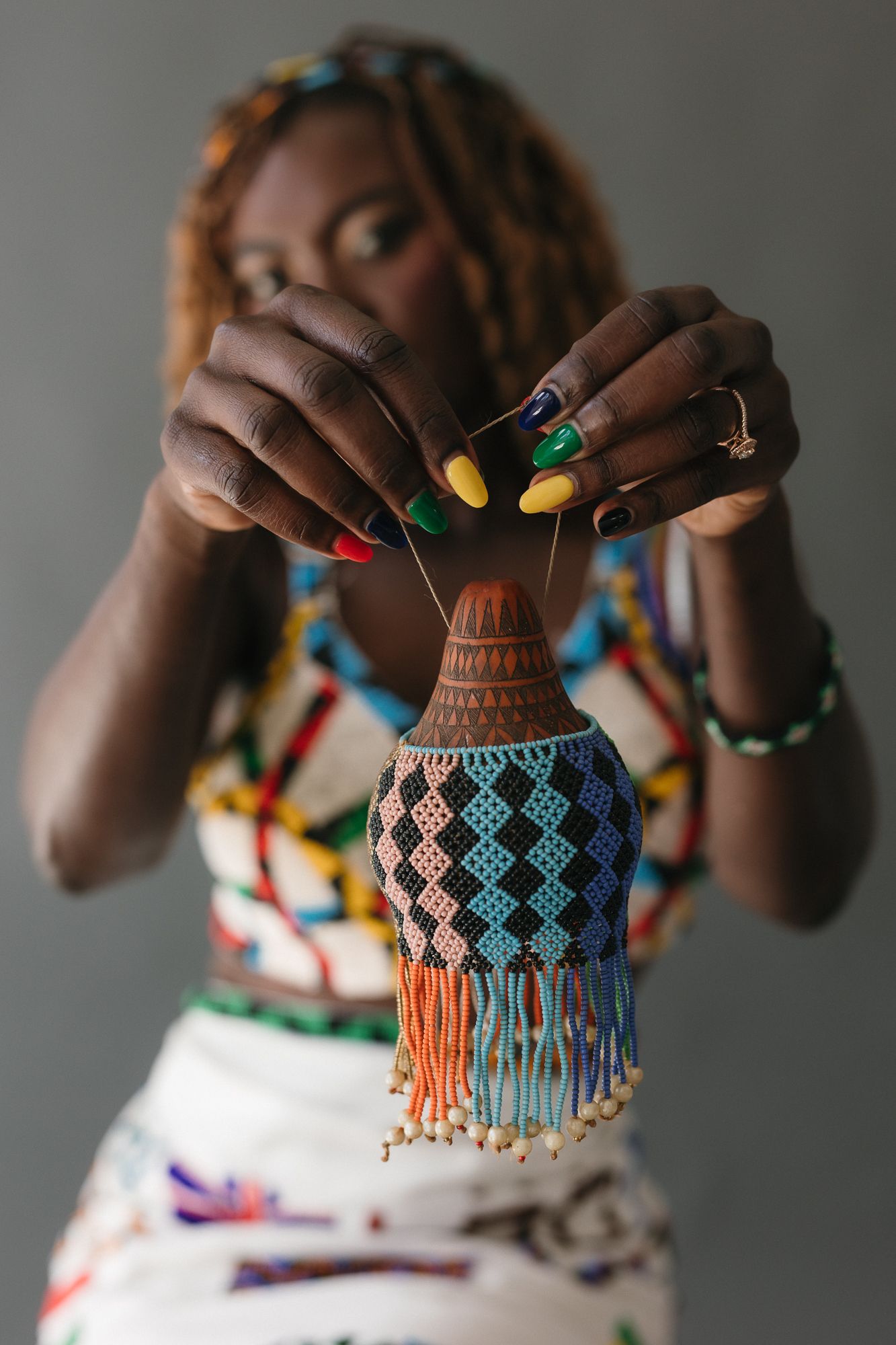 African woman with colorful nails holding a decorative beaded vase with fringe, wearing traditional patterned clothing.