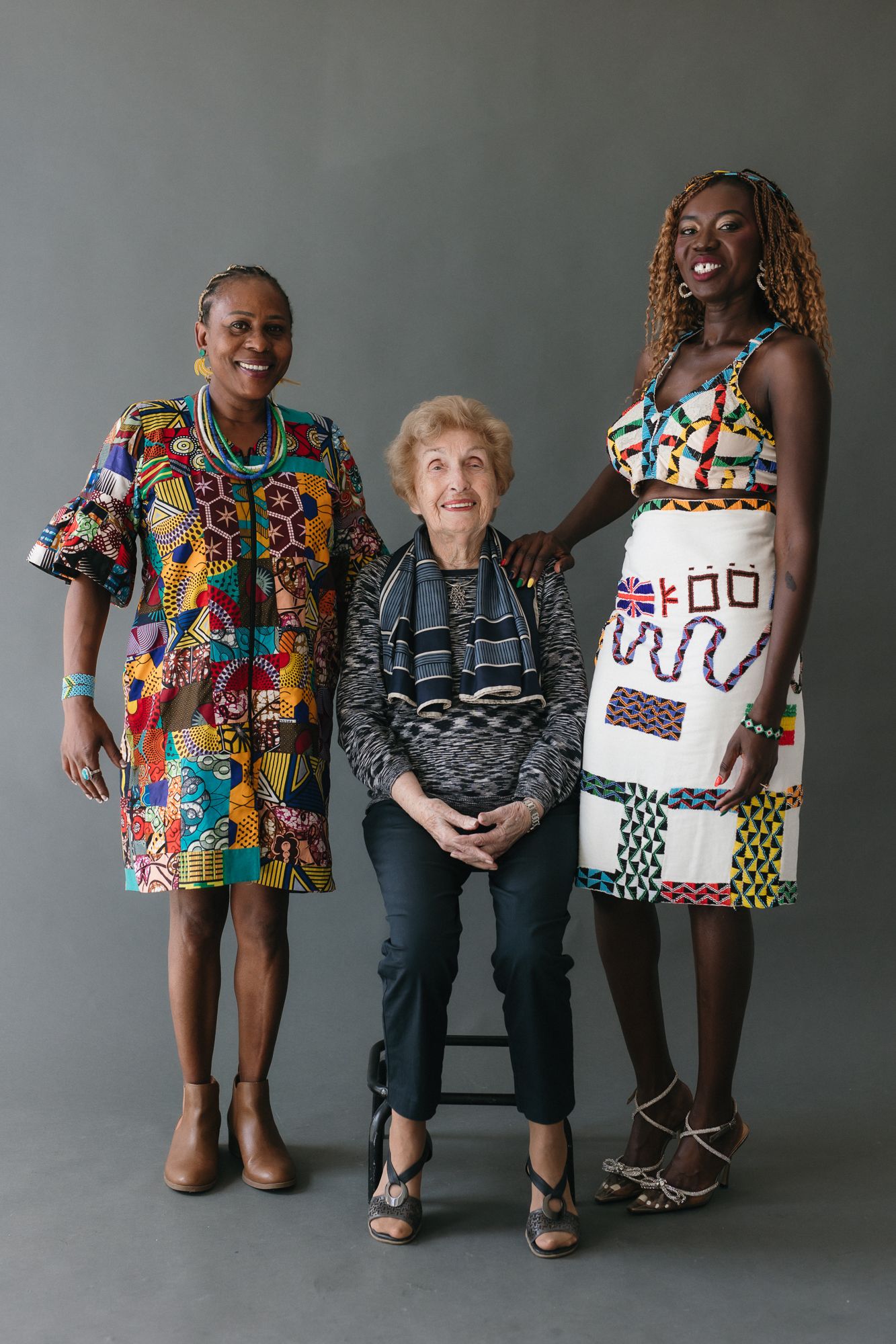 Three women posing together, two in colorful African print dresses flanking an elderly woman seated on a stool.
