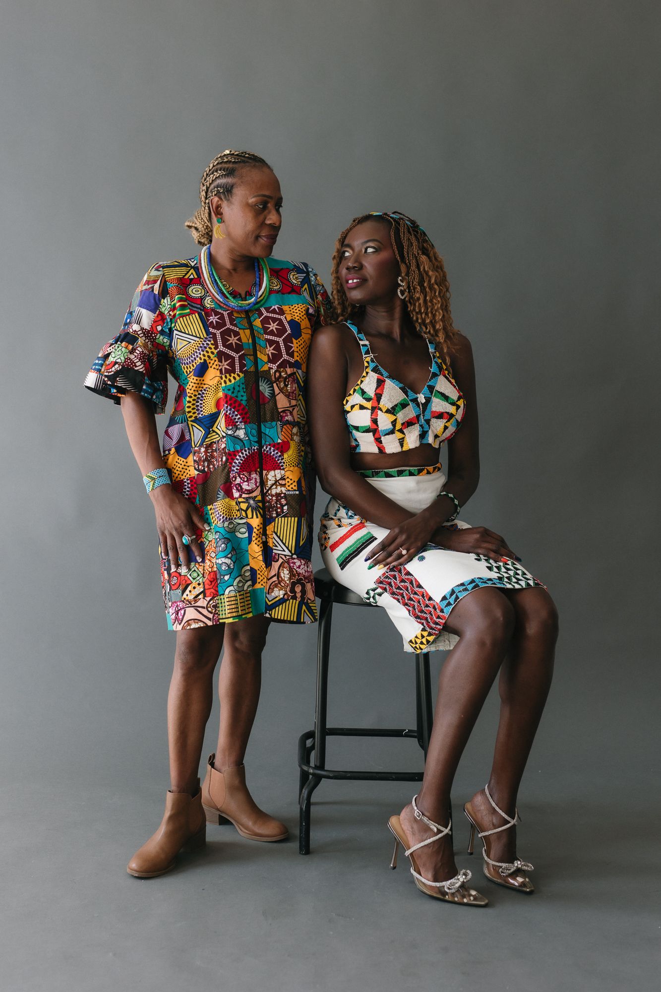 Two women in vibrant African print dresses pose together in a studio, one standing and one seated on a stool.