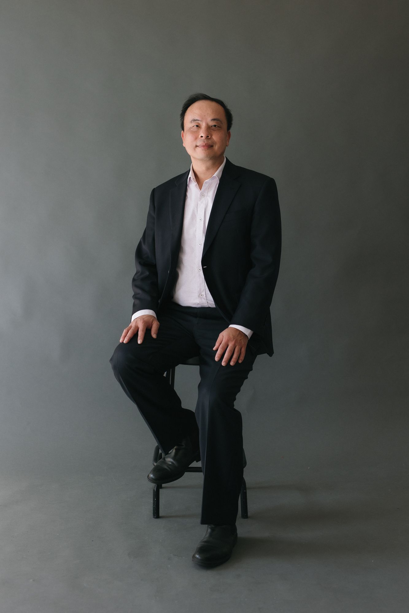 Middle-aged Asian man in black suit sitting on stool against gray background in professional portrait photo.